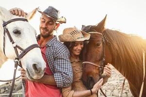 Happy couple having fun with horses inside stable - Young farmers sharing time with animals in corral ranch - Human and animals lifestyle concept photo