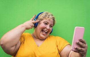 Happy plus size woman taking selfie with mobile smartphone and listening music with wireless headphones - Confident overweight people and technology concept photo