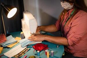 Hispanic woman working with sewing machine doing homemade face mask for preventing and stop corona virus spreading - Textile seamstress and homemade people work concept photo