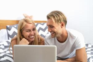 Happy couple lying on bed while using laptop computer - Happy people having funny movie bed time - Love relationship and technology concept photo