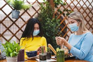 Multiracial women using mobile smartphone while having healthy lunch in coffee brunch bar during corona virus outbreak - Food and technology concept photo