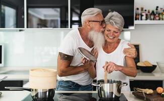 Happy senior couple having fun cooking together at home - Elderly people preparing health lunch in modern kitchen - Retired lifestyle family time and food nutrition concept photo