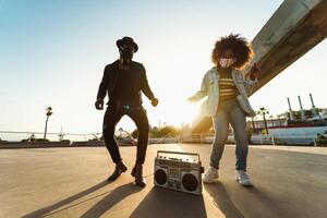 joven afro amigos vistiendo cara máscara bailando al aire libre mientras escuchando a música con inalámbrico auriculares y Clásico boombox foto