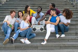 Young multiracial friends having fun listening music with vintage boombox stereo and using mobile smartphone while sitting on urban stairs - Youth millennial lifestyle concept photo