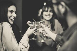 Happy young friends toasting and cheering cocktails at disco bar - Multiracial people having fun enjoying drinks at club - Black and white editing - Focus on glasses photo