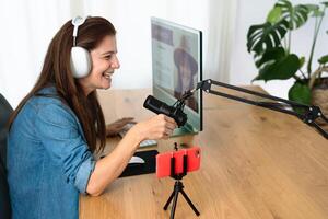 Mature woman recording a podcast using microphone and laptop while streaming on social network with mobile smartphone from her home studio photo