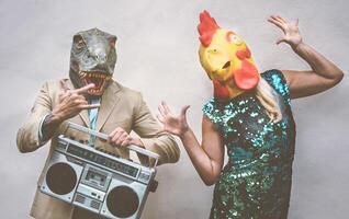 Crazy senior couple wearing chicken and t-rex mask while dancing outdoor - Mature trendy people having fun celebrating and listening music with boombox - Absurd concept of masquerade funny holidays photo