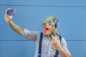 Crazy senior man wearing t-rex mask while taking selfie with mobile smartphone -  Hipster male having fun listening music and dancing outdoor -  Absurd surreal and technology concept photo