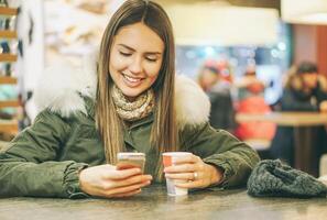 joven hermosa mujer Bebiendo un café en un café bar mientras mecanografía en móvil inteligente teléfono utilizando solicitud charla - bonito niña disfrutando un caliente bebida acecho social red en teléfono - Clásico filtrar foto