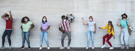 Happy friends wearing face mask listening music with vintage boombox outdoor - Multiracial young people having fun dancing together during corona virus outbreak - Youth millennial friendship concept photo