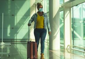 African senior woman walking in airport terminal with luggage wearing face mask during coronavirus pandemic photo