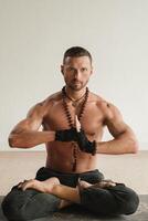 un hombre con un desnudo torso es sentado en el loto posición antes de haciendo yoga adentro. aptitud entrenador foto