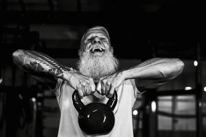 Senior fitness man doing kettle bell exercises inside gym - Fit mature male training in wellness club center - Body building and sport healthy lifestyle concept - Black and white editing photo