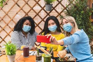 Multiracial friends wearing face mask while taking selfie with mobile smartphone cam in coffee brunch restaurant during corona virus outbreak photo