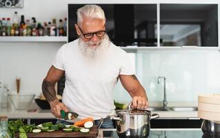 Happy senior man having fun cooking at home - Elderly person preparing health lunch in modern kitchen - Retired lifestyle time and food nutrition concept photo