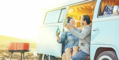 Travel couple having fun drinking beers sitting on their vintage camper mini van - Happy people with their pet enjoying barbecue at sunset during a roadtrip -  Concept of vacation lifestyle and love photo