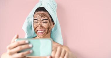 joven sonriente mujer tomando selfie mientras haciendo café fregar facial máscara - contento niña teniendo piel cuidado spa día a hogar - sano alternativa natural exfoliación tratamiento y tecnología concepto foto