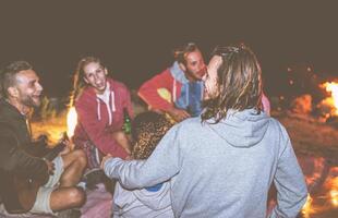 grupo de amigos teniendo divertido jugando guitarra siguiente a el hoguera a noche - contento joven personas cámping juntos riendo y Bebiendo cerveza - amistad, vacaciones, Días festivos concepto foto