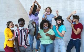 Happy friends wearing face mask listening music with vintage boombox outdoor - Multiracial young people having fun dancing together during corona virus outbreak - Youth millennial friendship concept photo