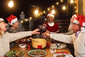 Happy multiracial senior friends toasting with red wine glasses during Christmas holidays dinner celebration on patio house party photo