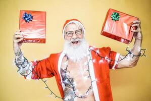 Happy tattoo hipster santa claus equiped with white lights giving christmas gifts - Trendy beard senior wearing xmas clothes and holding presents - Celebration and holidays concept photo