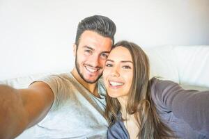 Happy young couple taking a selfie with mobile smart phone camera in the living room embracing on sofa at home - Friends making self portrait on the couch - People, relationship, lifestyle concept photo
