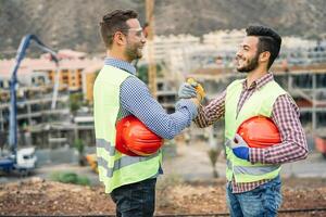 Young builders shaking hands making an agreement on costruction site - Workers reaching a deal and laughing - Building, dealing, engineer industrial concept photo