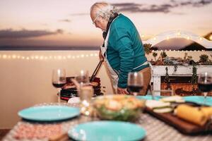 mayor hombre interrogatorio intenso carne a parilla cena en terraza - abuelo cocinando para su familia en techo - concepto de comida barbacoa y comiendo juntos al aire libre foto