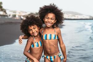 Happy sisters enjoying inside sea water during summer time - Afro kids having fun playing on the beach - Family love and travel vacation lifestyle concept photo