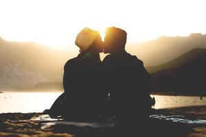 romántico joven Pareja besos en el playa en puesta de sol - silueta de adolescentes amantes a el comenzando de su historia sentado en arena - gente, amar, estilo de vida, relación concepto foto