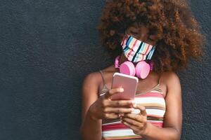 Happy Afro woman using mobile smartphone while wearing face colored mask outdoor photo