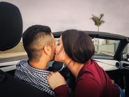 Young couple kiss in a convertible car during their road trip - Happy romantic newlywed date driving a cabriolet auto in honeymoon - Love, relationship and youth holidays lifestyle photo