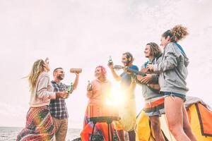 Group of friends having barbecue drinking beers while camping on the beach - Happy people enjoying camp bbq playing guitar, singing and listening music at sunset -  Friendship, adventure concept photo