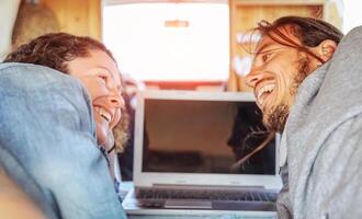 Happy man and woman lookin each other and using their computer in caravan - Travel couple tender moment during their journey on vintage minivan with wood interior - Vacation, love, technology concept photo