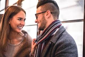 Happy couple laughing while looking each other in the bus - Young beautiful woman pulling her boyfriend by scarf next to her - Love, romantic, relationship concept - Focus on man eye photo