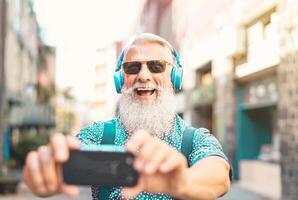 Senior bearded man taking selfie with mobile phone while listening favorite playlist with headphones - Trendy hipster male having fun with smartphone social apps outdoor - People technology concept photo