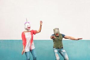 Happy crazy couple dancing while wearing t-rex and unicorn mask outdoor - Seniors having fun celebrating carnival fest - People culture lifestyle and absurd disguise concept photo