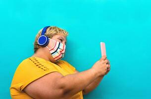 Plus size woman taking selfie with mobile smartphone and listening to music with wireless headphones while wearing face protective mask to avoid corona virus infection - Health and technology concept photo