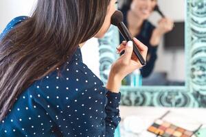 Young woman applying makeup while looking in mirror - Happy girl using make up brush - Cosmetic and lifestyle people concept photo