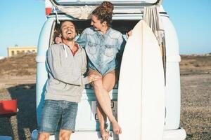 Happy surfers couple standing behind on vintage camper mini van - Young people adventuring on road trip with a minivan transport - Concept of travel, vacation, relationship and youth lifestyle photo