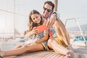 Father and daughter taking selfie smartphone camera during sailboat trip - Happy family having fun with new mobile app for social media on vacation - Travel people lifestyle and technology concept photo