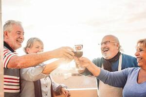 contento personas mayores amigos Bebiendo rojo vino en terraza puesta de sol - pensionistas personas teniendo divertido disfrutando hora juntos al aire libre - mayor Jubilación estilo de vida concepto foto