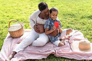 Happy African pregnant mother spending time with his son doing a picnic during weekend in public park - Afro family watching on smartphone - Maternity and parents lifestyle concept photo