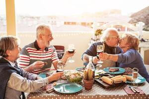 Happy senior friends dining and cheering with red wine at barbecue in terrace outdoor - Retired people having fun at bbq party on patio - Friendship, food and elderly lifestyle concept photo