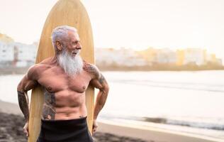 Senior male having fun surfing during sunset time - Fit retired man training with surfboard on the beach - Elderly healthy people lifestyle and extreme sport concept photo