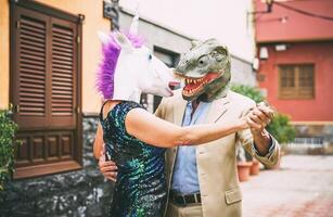 Crazy couple dancing and wearing dinosaur t-rex and unicorn mask - Senior elegant people having fun masked at carnival parade - Absurd, eccentric, surreal, fest and funny masquerade concept photo