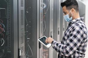 Male informatic engineer working inside server room database while wearing face mask during corona virus outbreak photo
