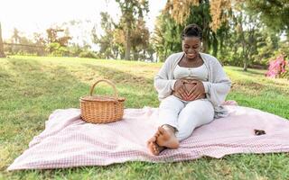Happy Afro woman touching her pregnant belly doing heart shape with hands in public park - Maternity lifestyle concept photo