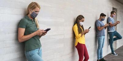 Young people wearing face mask using mobile smartphone outdoor - Multiracial friends having fun with new technology social media app during corona virus outbreak - Youth millennial lifestyle concept photo