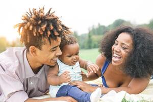 contento africano familia disfrutando un picnic soleado día al aire libre - madre y padre teniendo divertido con su hija en un público parque - amar, padres y felicidad concepto foto